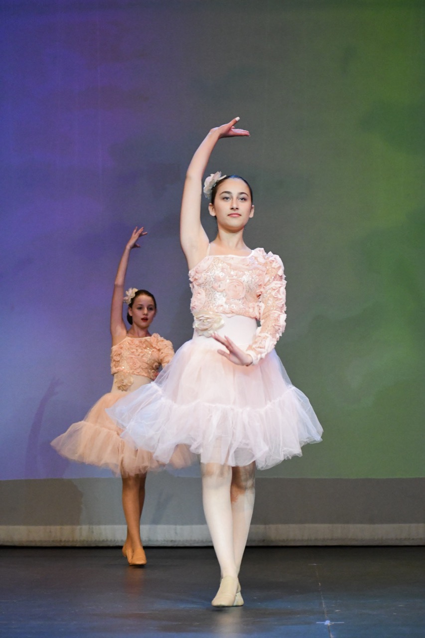 Two dancers in peach costumes performing on stage, with the foreground dancer raising one arm.